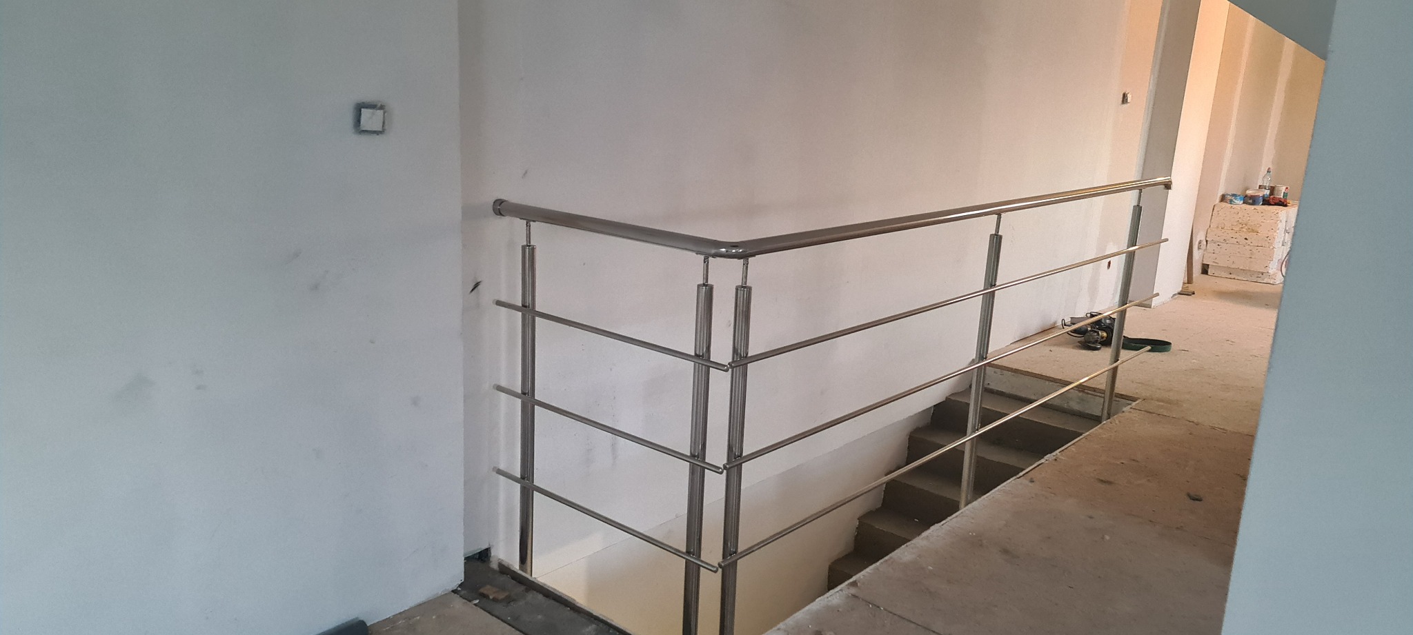 Nowoczesna, nierdzewna balustrada schodowa z czterema poziomami poprzeczek, zamontowana w surowym wnętrzu w trakcie remontu, widoczne przewody elektryczne i materiały budowlane w tle.