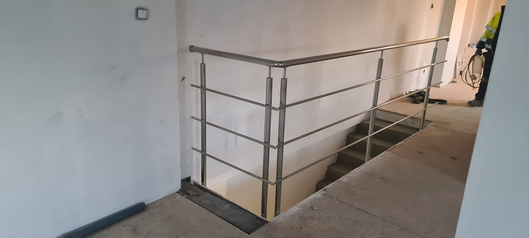 Nowoczesna balustrada ze stali nierdzewnej zamontowana przy schodach w surowym stanie, widoczny fragment pracownika w odblaskowej kamizelce.