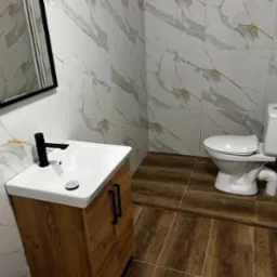 Wykończona łazienka z marmurowymi płytkami na ścianach, drewnopodobną podłogą, białą umywalką z czarną armaturą i toaletą.