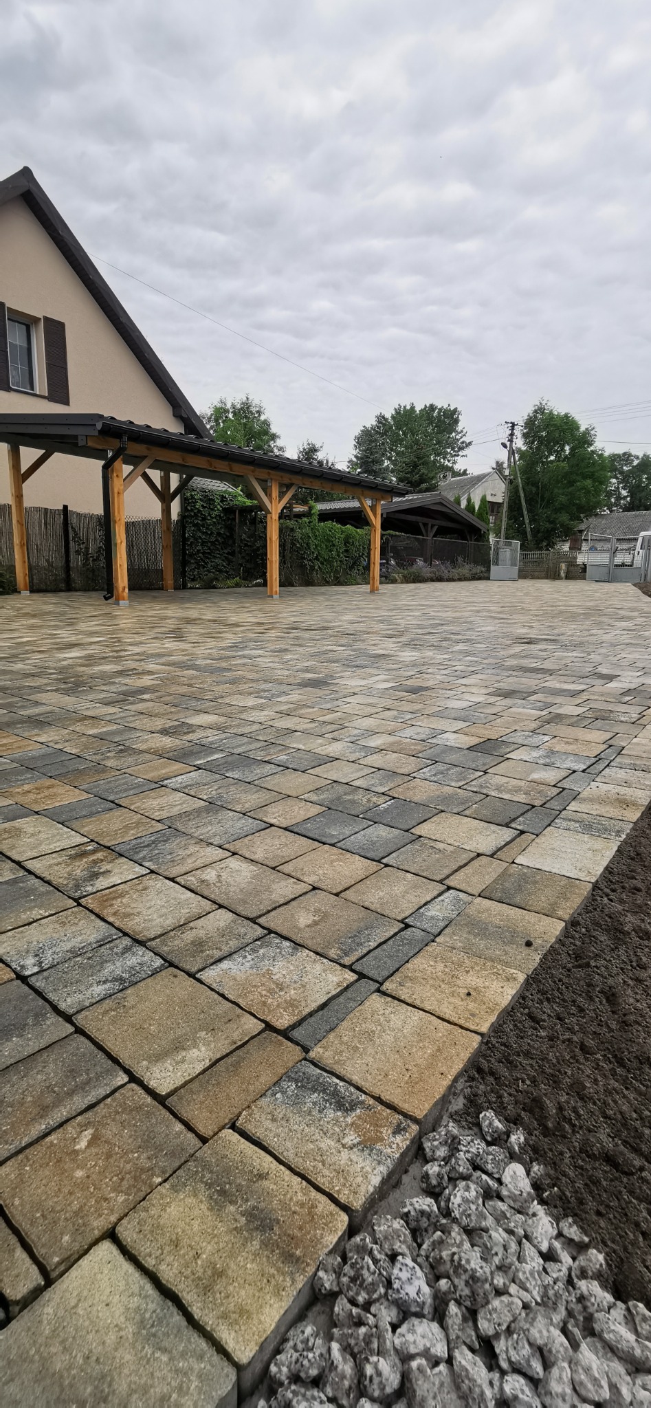 Ułożona, wielobarwna kostka brukowa na podjeździe z drewnianą wiatą garażową w tle, widoczne obrzeża z kamieni.