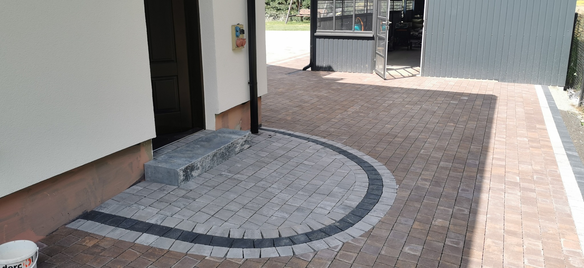 Ułożona kostka brukowa w dwóch kolorach (szarym i brązowym) tworząca półokrąg przed wejściem do domu, z widocznym stopniem i puszką elektryczną na ścianie budynku.