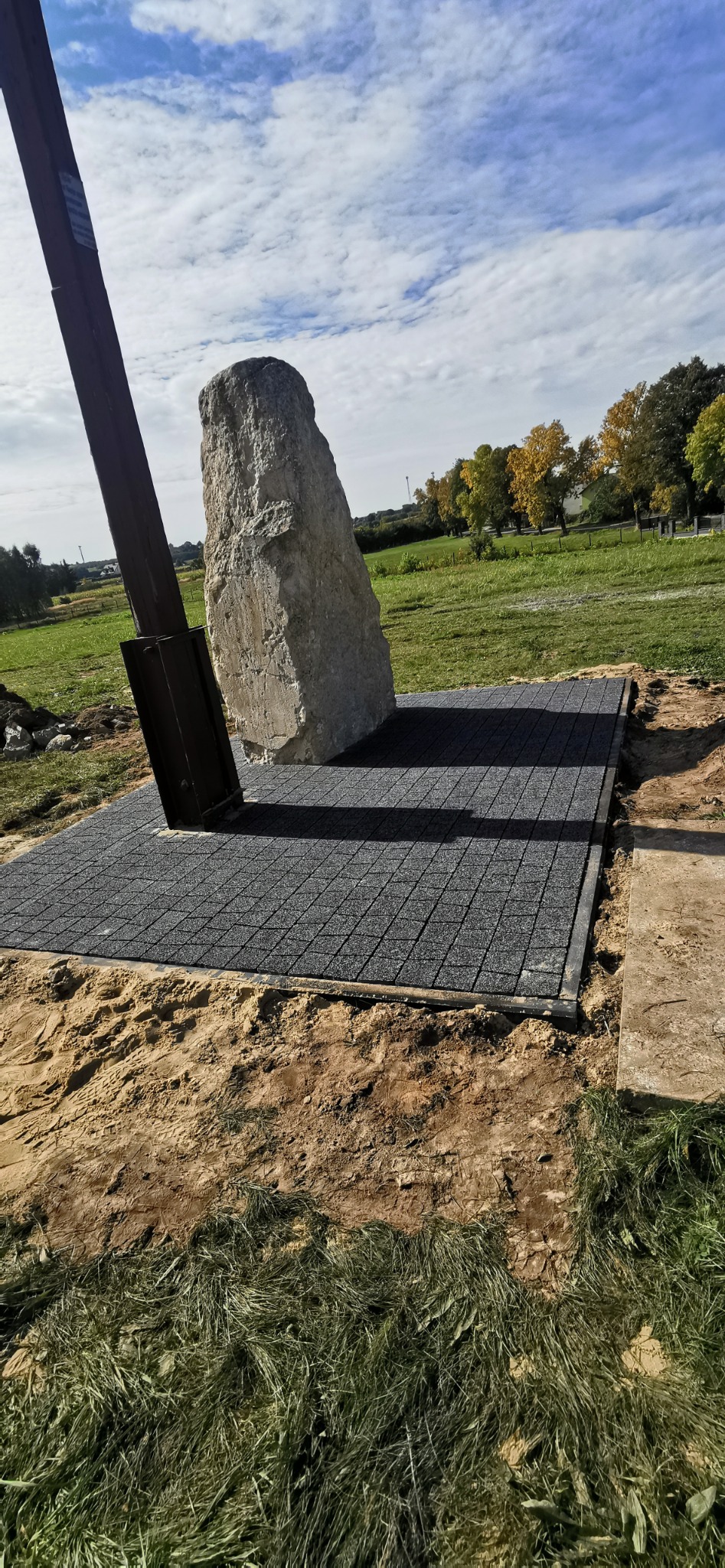 Monumentalny kamień wsparty na metalowej konstrukcji, stojący na nowo ułożonej, szarej nawierzchni z kostki, otoczony ziemią i trawą, na tle błękitnego nieba z chmurami.