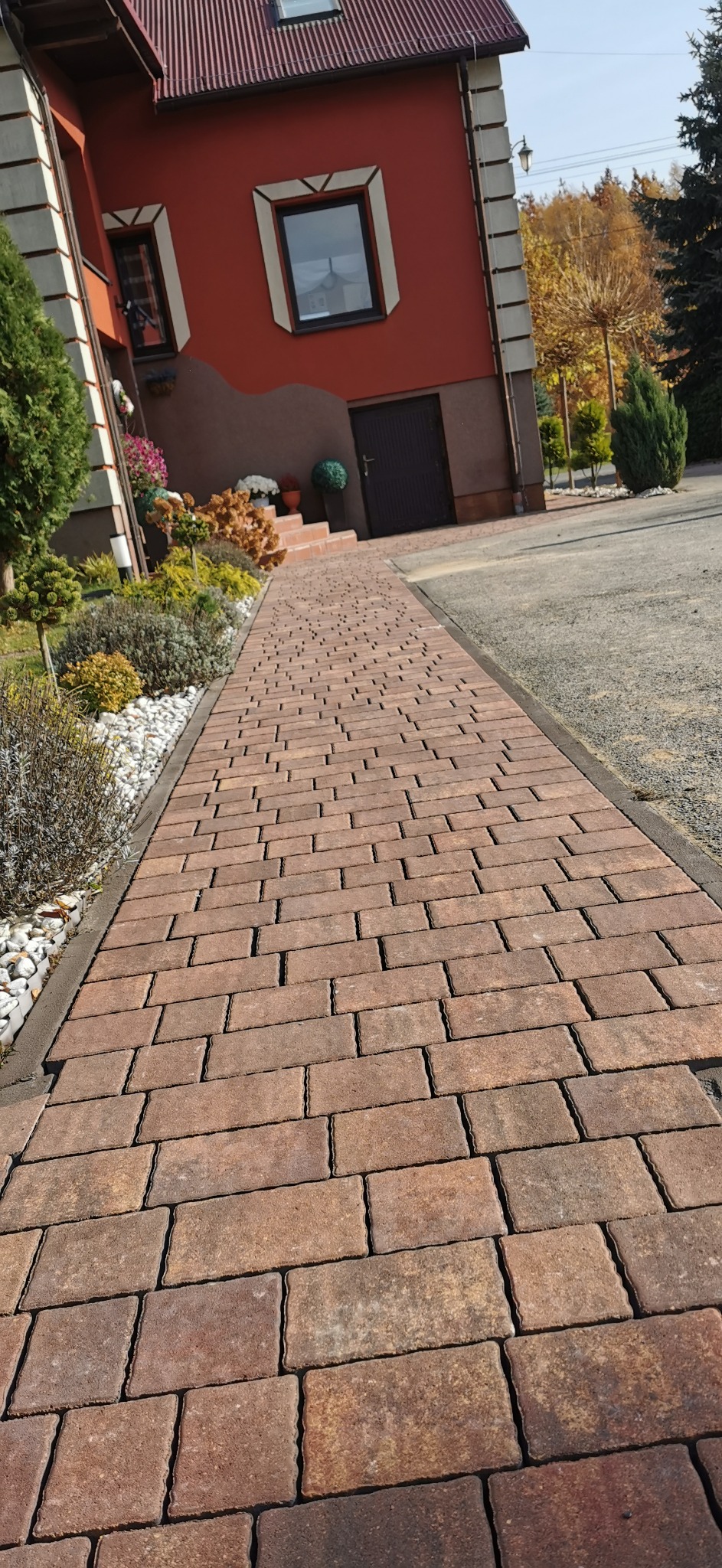 Ścieżka z brązowej kostki prowadząca do domu z czerwonym dachem, obok rabata z białymi kamieniami i roślinami.