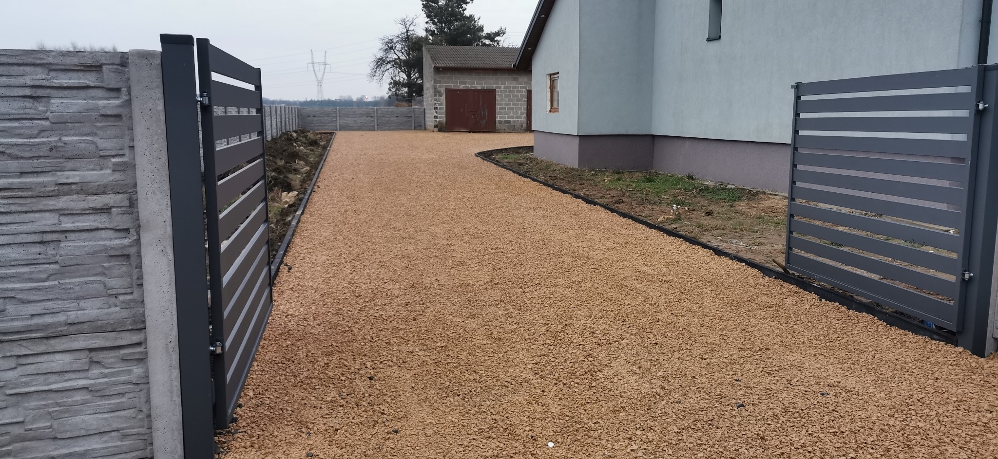 Podjazd wysypany drobnym, brązowym kruszywem, ograniczony czarnym obrzeżem, z szarą, przesuwną bramą panelową i fragmentem ogrodzenia z szarego betonu imitującego kamień. W tle budynek gospodarczy...