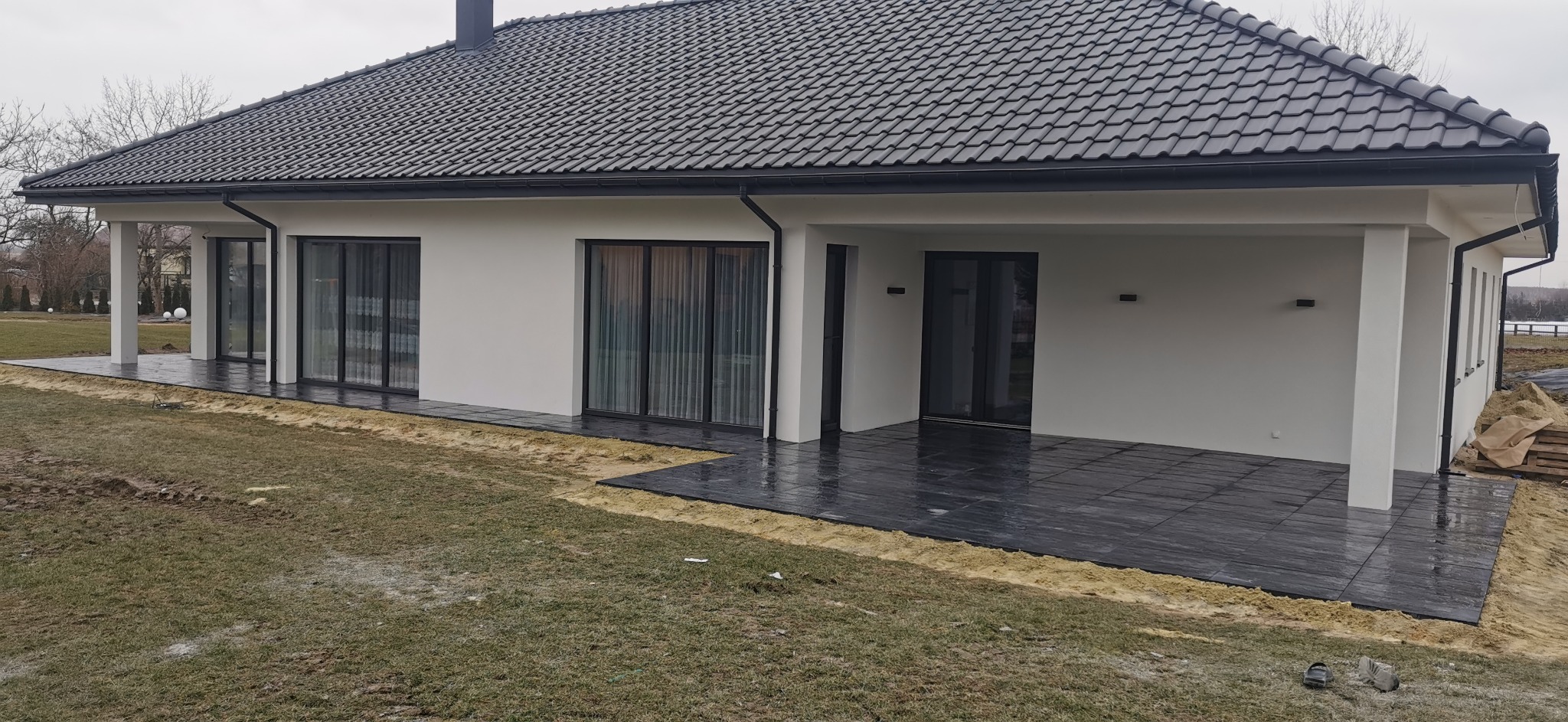 Nowoczesny dom w Częstochowie z wykończonym tarasem z ciemnej kostki brukowej, widoczne duże przesuwne okna i fragment trawnika.