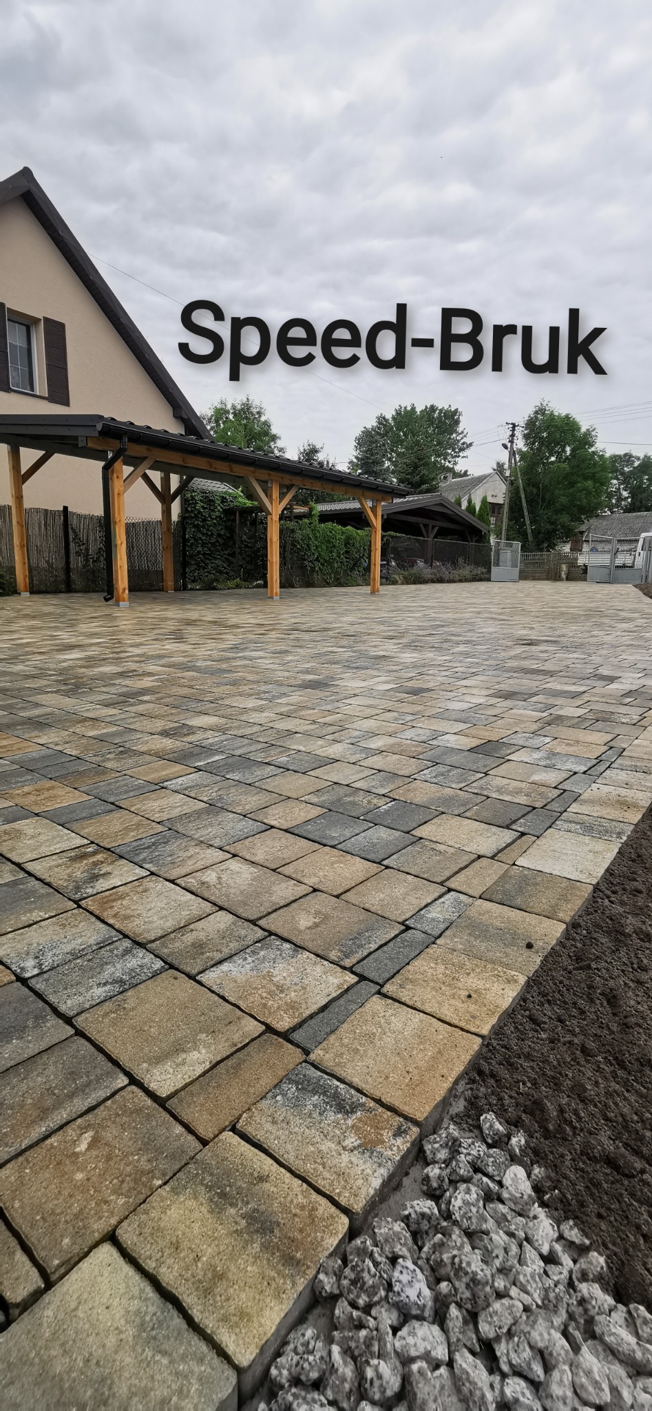 Ułożona, wielokolorowa kostka brukowa na podjeździe przed domem z drewnianą wiatą, widoczne obrzeża z kamieni i świeżo obsypana ziemia.