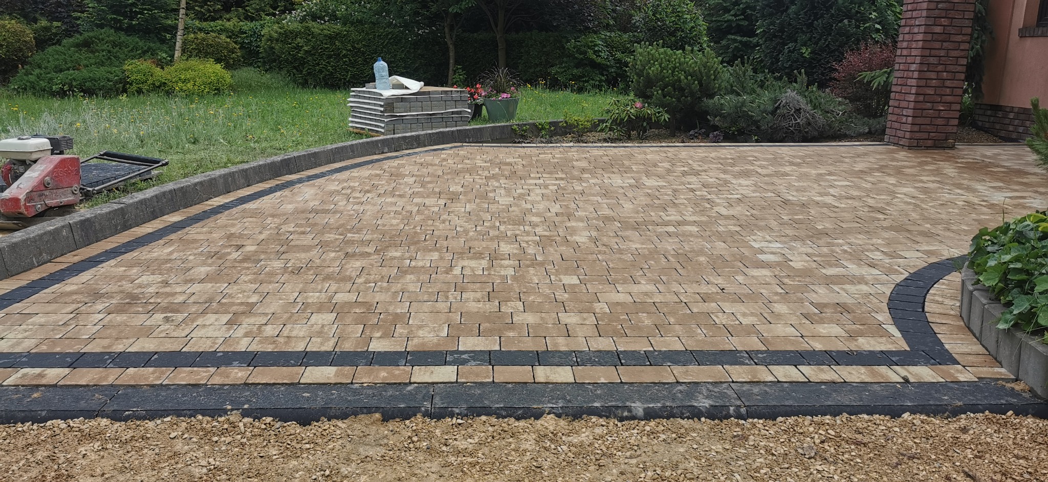 Ułożona beżowo-brązowa kostka na podjeździe z obramowaniem z ciemnoszarej kostki, widoczna ubijarka wibracyjna, paleta z kostką i butelka wody w tle.