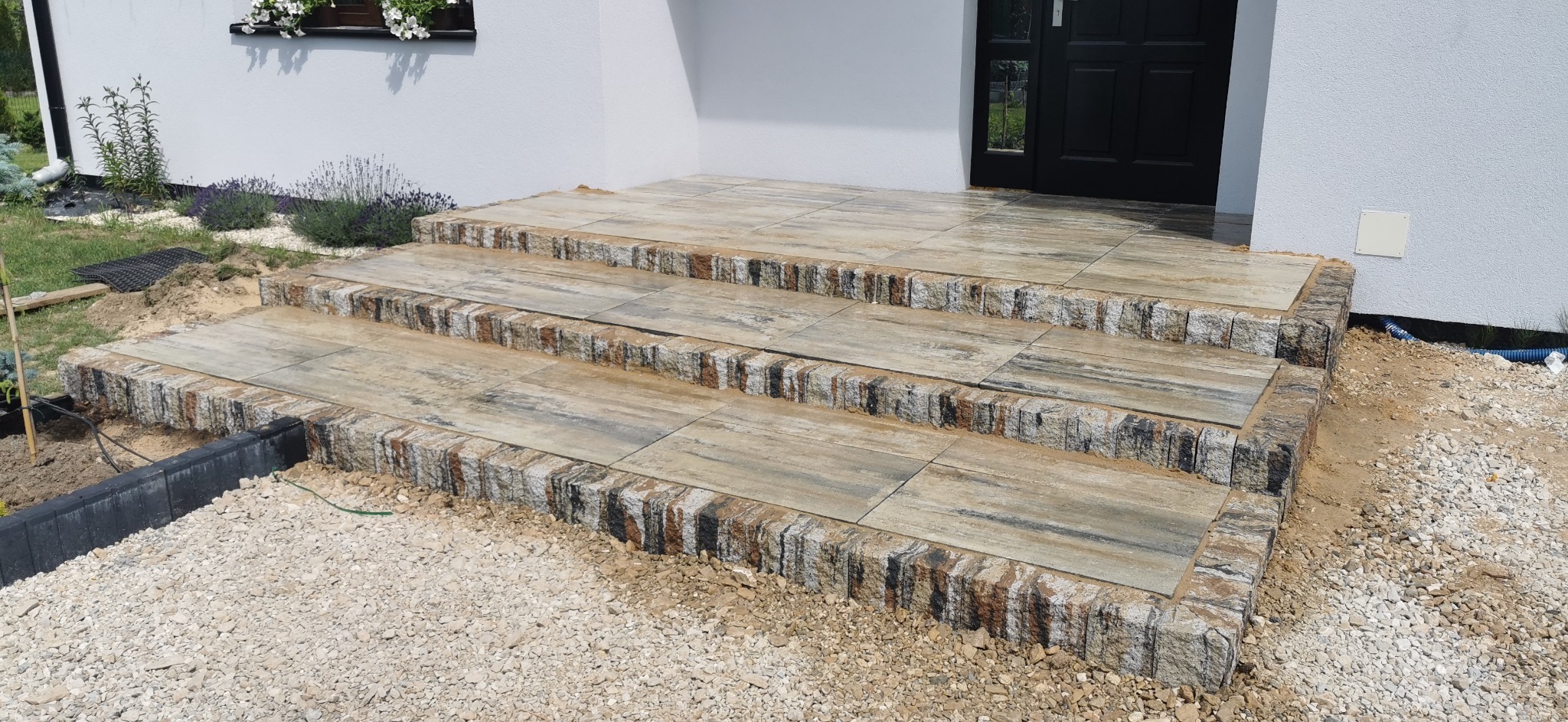 Trzy stopniowe schody wejściowe wykończone płytami imitującymi drewno i obrzeżami z kostki o nieregularnej fakturze, otoczone żwirem i fragmentem ogrodu.