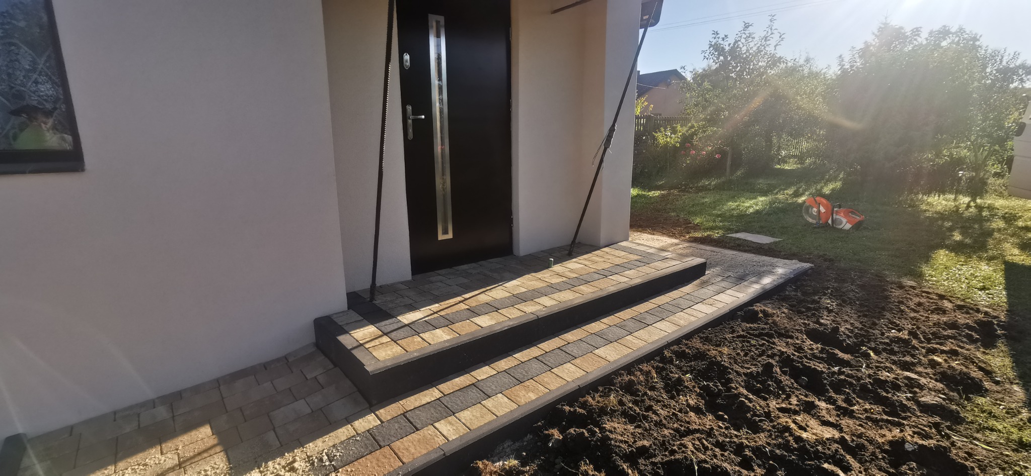 Ułożone schody z kostki brukowej w odcieniach szarości i beżu prowadzące do czarnych drzwi wejściowych, z widocznym narzędziem do cięcia kostki na trawniku w tle.