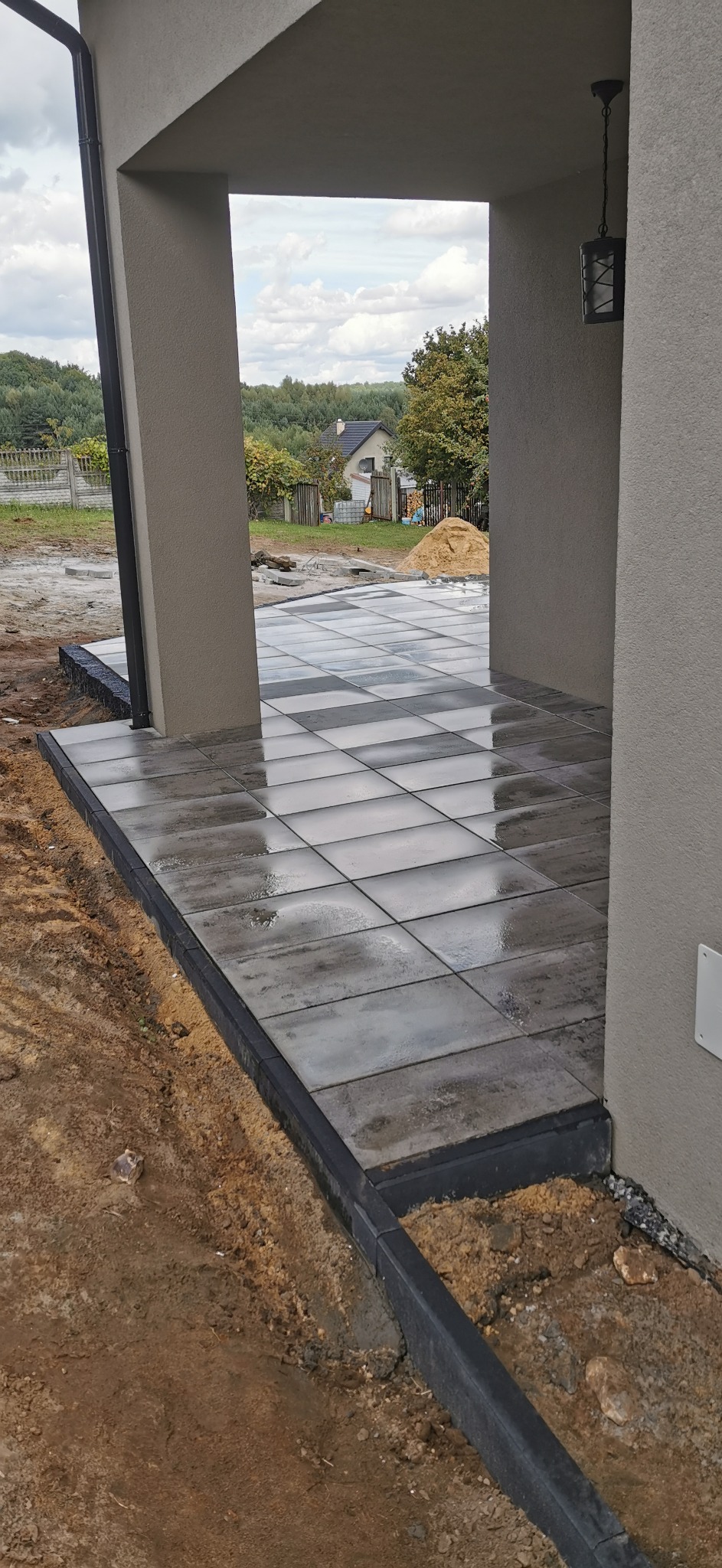 Świeżo ułożona, mokra, szara kostka na tarasie z czarnym obrzeżem, widok na ogród i drzewa w tle, lampa na suficie tarasu.