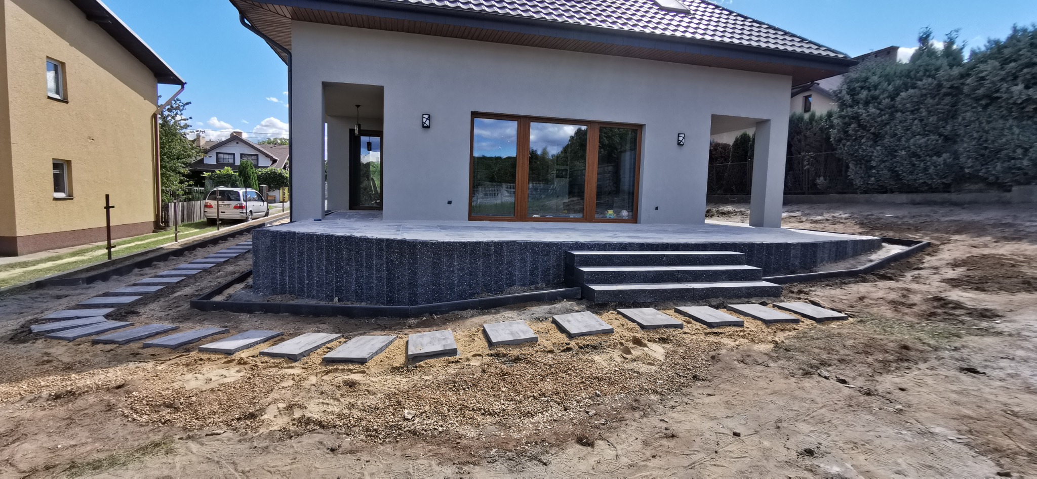 Nowoczesny taras z szarej kostki brukowej, schody i ścieżka z płyt na podsypce piaskowej, dom jednorodzinny w tle.