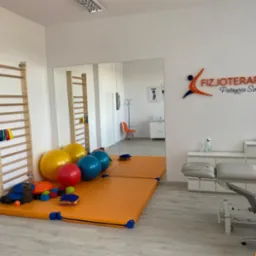 Jasne wnętrze gabinetu z drabinkami gimnastycznymi, materacami, piłkami do ćwiczeń oraz stołem do masażu i logo 'Fizjoterapia Patrycja Sznaza' na ścianie.