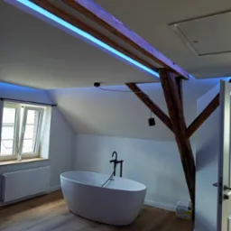 Jasna łazienka na poddaszu z drewnianymi belkami oświetlonymi niebieskim światłem LED, wolnostojąca wanna i drewniana podłoga. Nowoczesny design.