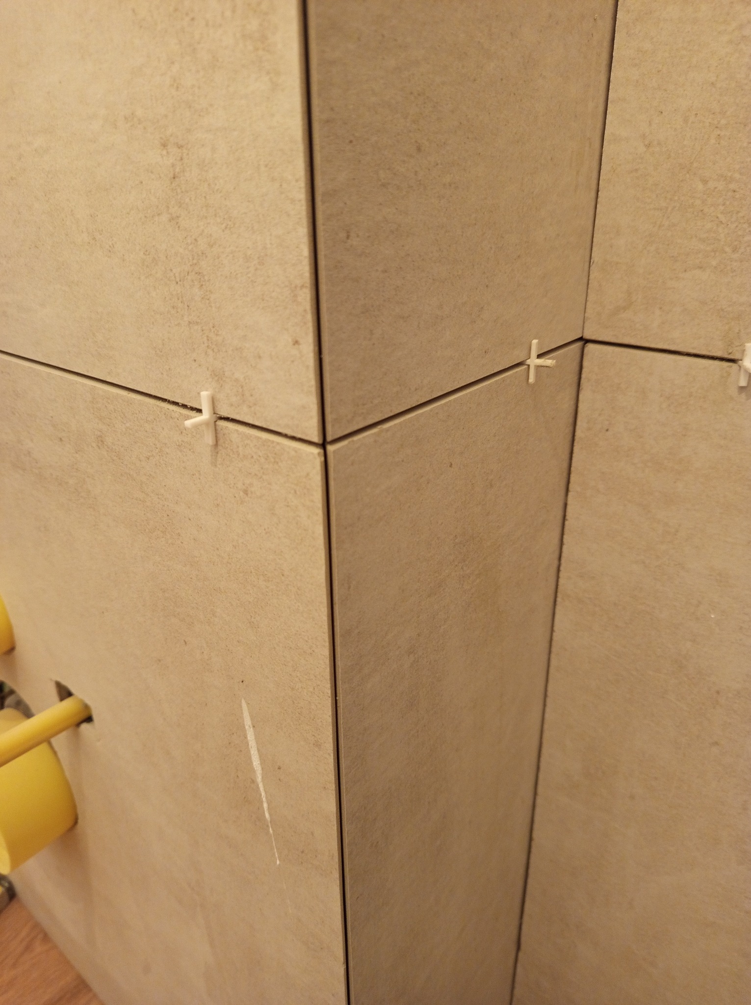 Beżowe płytki ścienne z krzyżykami dystansowymi, widoczne fugi i fragment instalacji. Ujęcie z bliska na narożnik ściany.