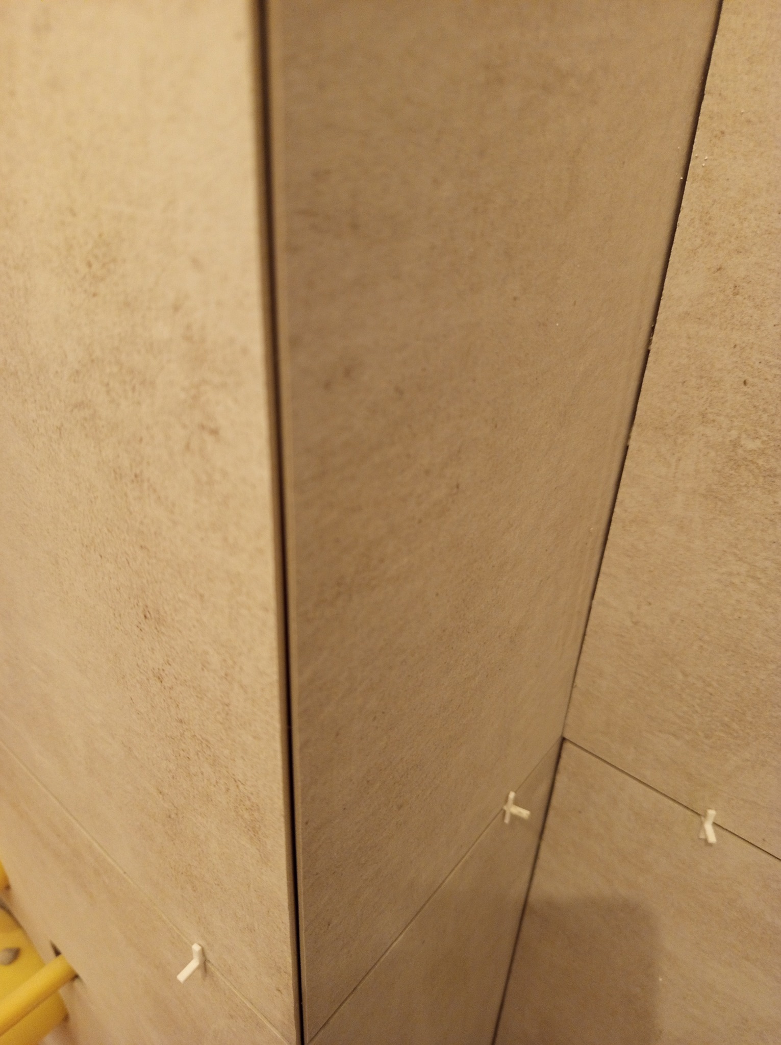 Beżowe płytki ścienne w trakcie montażu, krzyżyki dystansowe widoczne w spoinach. Układanie płytek w rogu pomieszczenia, zbliżenie na detale montażu.