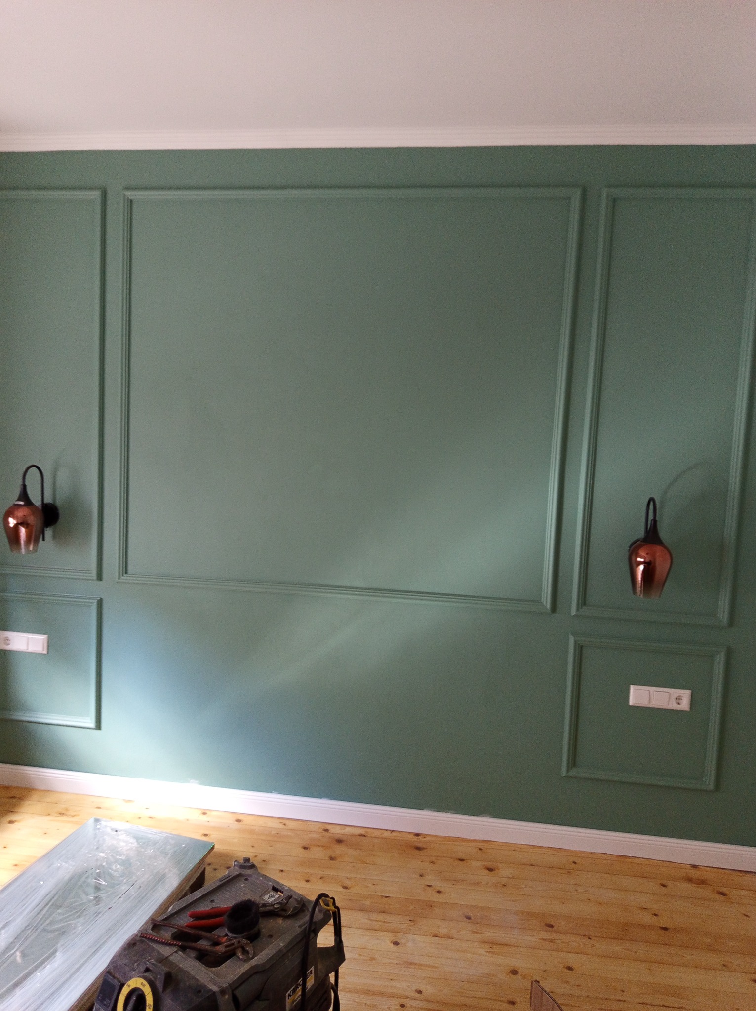 Oliwkowa ściana z dekoracyjnymi panelami i miedzianymi lampami. Na podłodze drewniane deski i narzędzia, sugerujące prace wykończeniowe w pomieszczeniu.