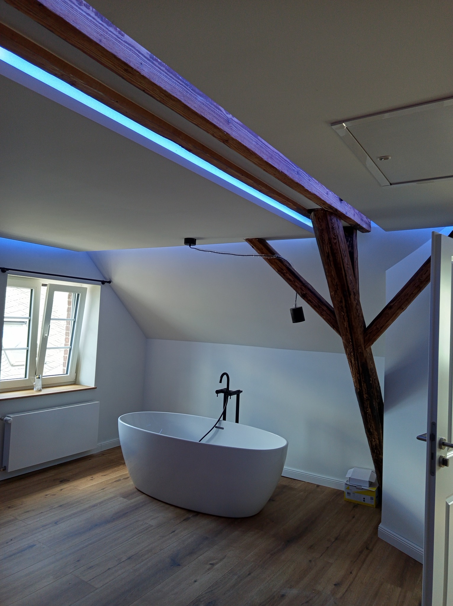 Jasna łazienka na poddaszu z drewnianymi belkami oświetlonymi niebieskim światłem LED, wolnostojąca wanna i drewniana podłoga. Nowoczesny design.