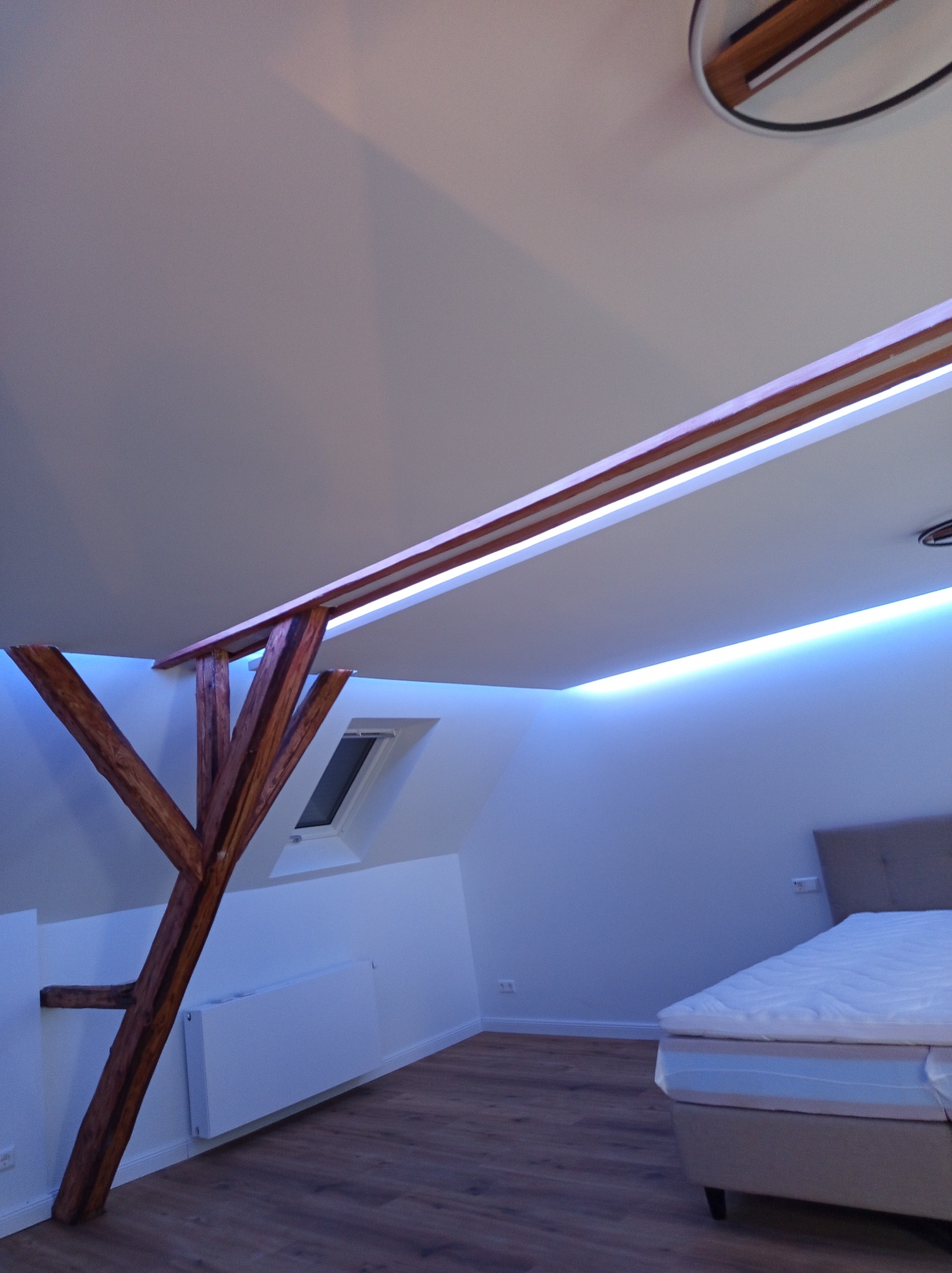 Pokój na poddaszu z drewnianą konstrukcją i listwami LED. Widoczne skosy, okno dachowe, łóżko i grzejnik. Oświetlenie tworzy specyficzny, chłodny klimat wnętrza.