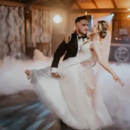 Para młoda w tańcu unoszona przez pana młodego na tle dekoracji sali weselnej z efektem mgły i światłami punktowymi.