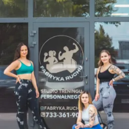 Trzy kobiety w sportowych strojach pozują przed wejściem do studia treningu personalnego Fabryka Ruchu z widocznym logo i numerem telefonu.