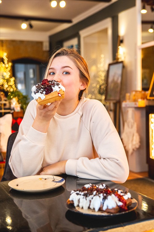 Młoda kobieta z piegami na policzkach, trzymająca w dłoni wafla z bitą śmietaną, polewą czekoladową i wiśniami, zbliżając go do ust, na stole talerz z resztkami wafla i drugi wafelek na talerzu,...