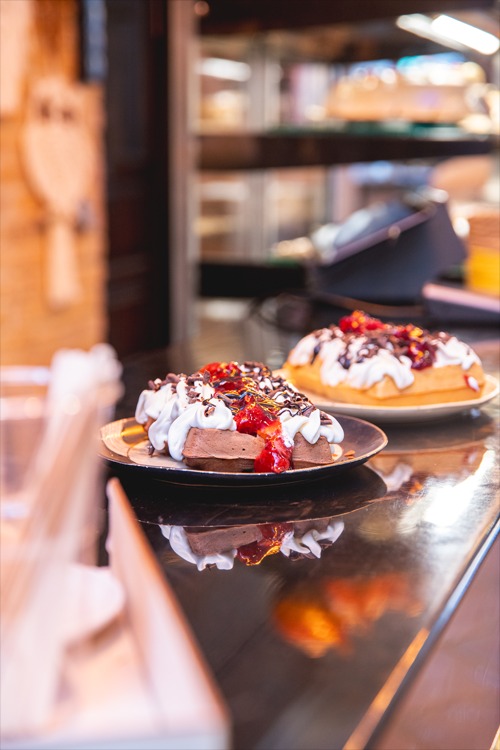 Dwa talerze z goframi: czekoladowy z bitą śmietaną, truskawkami i czekoladą, oraz jasny z bitą śmietaną i sosem truskawkowym, na tle lady chłodniczej.