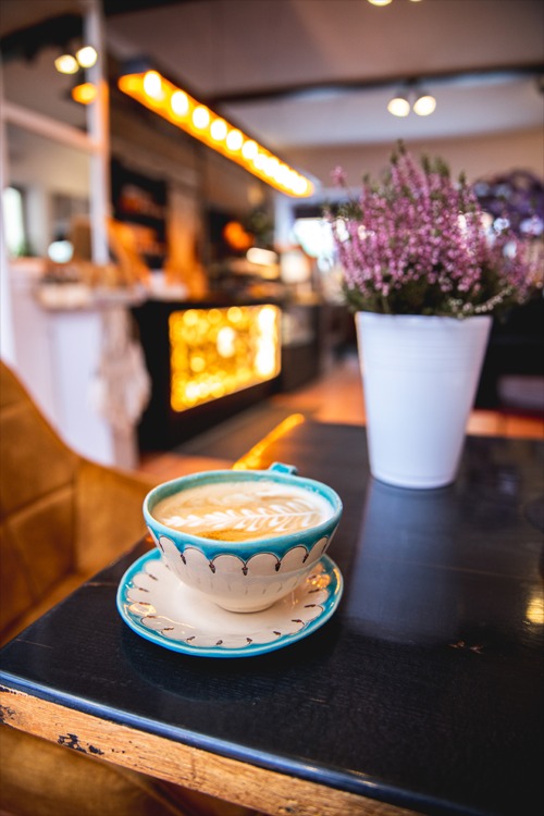 Kawa latte w ozdobnej filiżance z błękitnym wzorem na ciemnym drewnianym stoliku, w tle rozmyte wnętrze kawiarni z podświetlonym barem i doniczką z wrzosami.