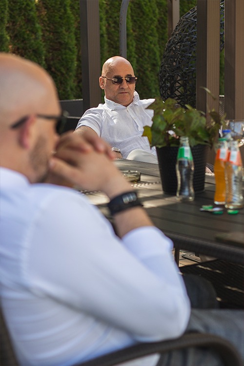 Dwaj mężczyźni siedzą przy stole na zewnątrz, jeden z nich w okularach przeciwsłonecznych i białej koszuli, drugi częściowo widoczny z przodu, również w białej koszuli, na stole napoje i roślina...