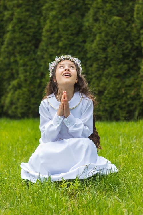 Dziewczynka w białej sukience i wianku klęczy na trawie z złożonymi rękami, spoglądając w górę na tle zielonych krzewów.