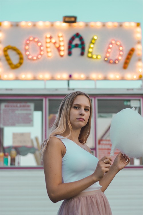 Młoda kobieta w białym topie i tiulowej spódnicy trzyma watę cukrową na tle budki z napisem 'Donald' oświetlonym żarówkami.
