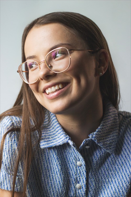 Portret uśmiechniętej młodej kobiety w okularach o złotych oprawkach, ubrana w niebieską tweedową koszulę z perłowymi guzikami, spoglądającej w lewo.