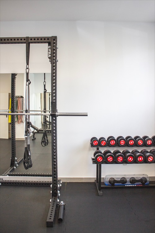 Domowa siłownia z czarną gumową podłogą, klatką do ćwiczeń, sztangą i stojakiem na hantle z czerwonymi akcentami.