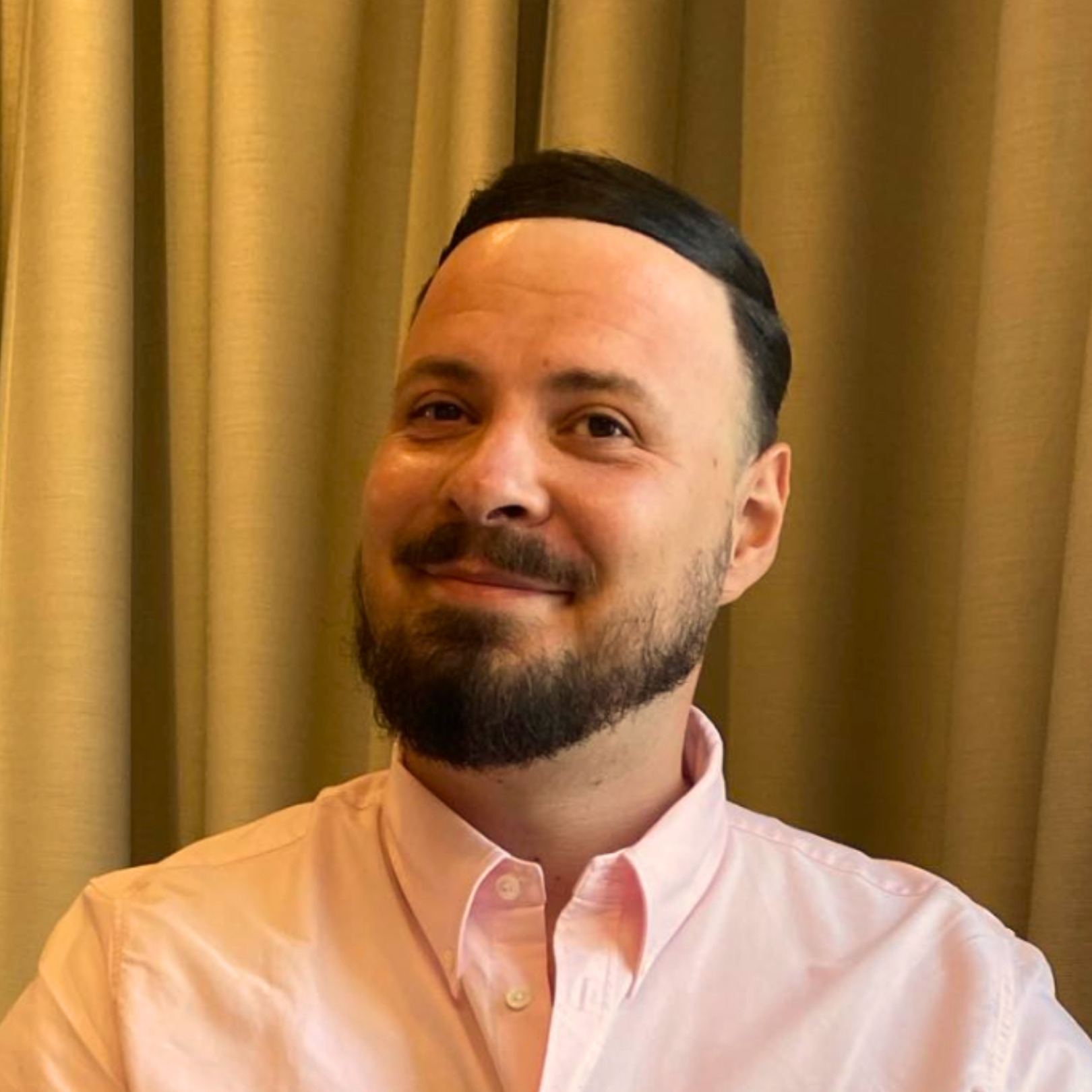 Portret uśmiechniętego mężczyzny z brodą w różowej koszuli na tle zasłon w odcieniach beżu.