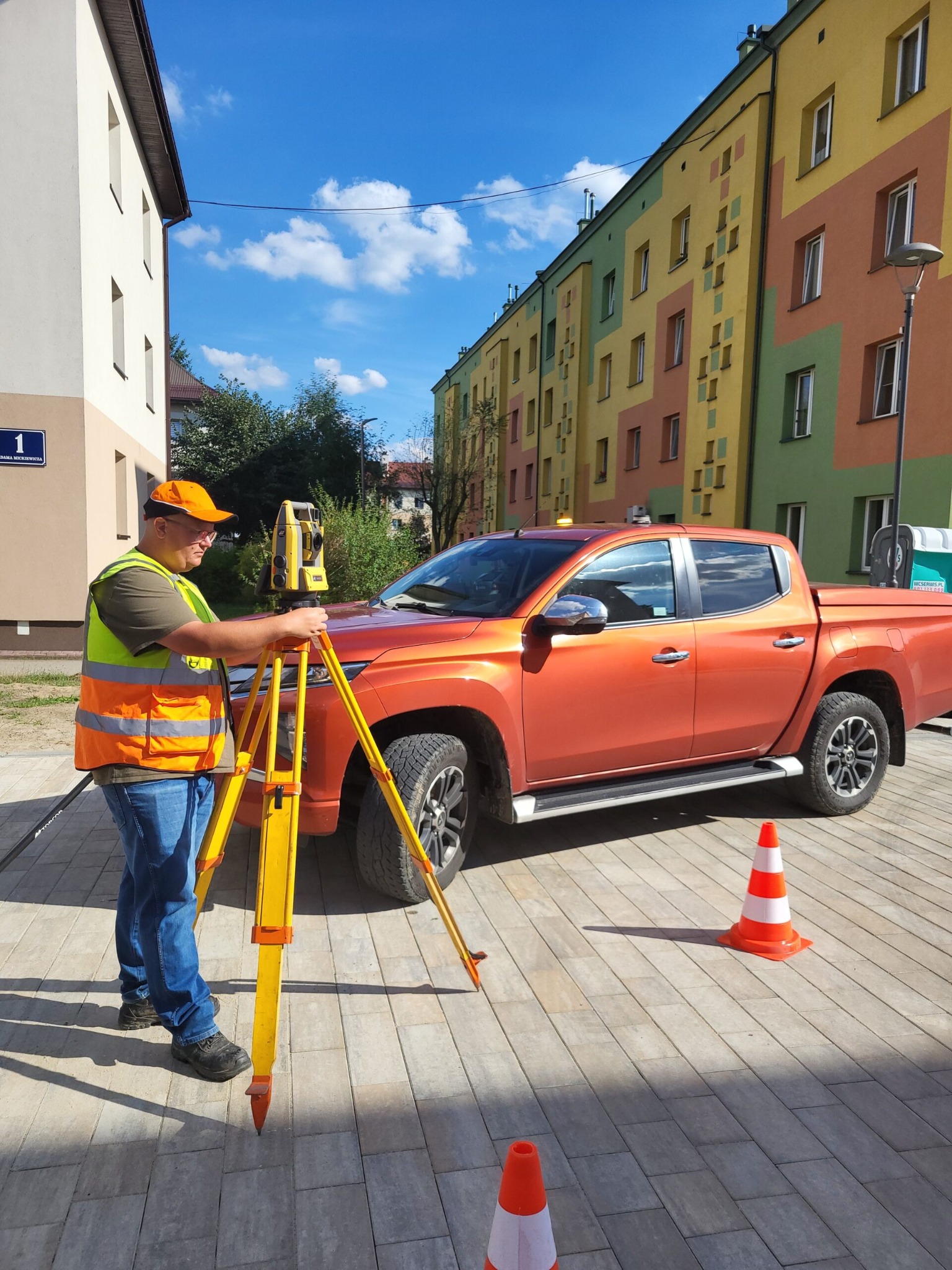 Mężczyzna w pomarańczowej kamizelce ustawia żółty niwelator na trójnogu przed pomarańczowym pickupem na tle kolorowego budynku.