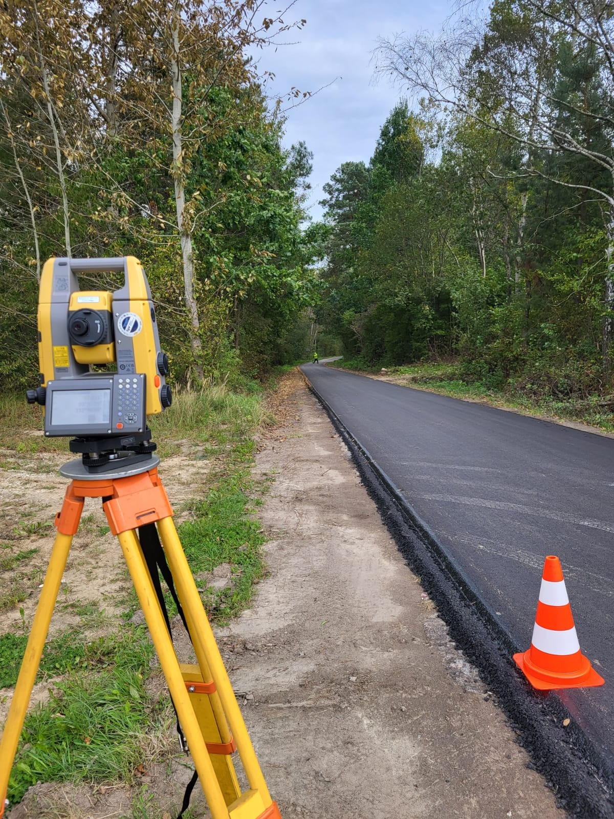 Pomiar geodezyjny powykonawczy nowej drogi asfaltowej w otoczeniu leśnym, z widocznym teodolitem na statywie i pachołkiem drogowym, w oddali osoba w jaskrawej kamizelce.