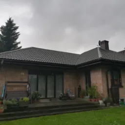 Dom jednorodzinny z brązową dachówką ceramiczną w pochmurny dzień, widoczna część elewacji z czerwonej cegły, roślinność w donicach na tarasie.