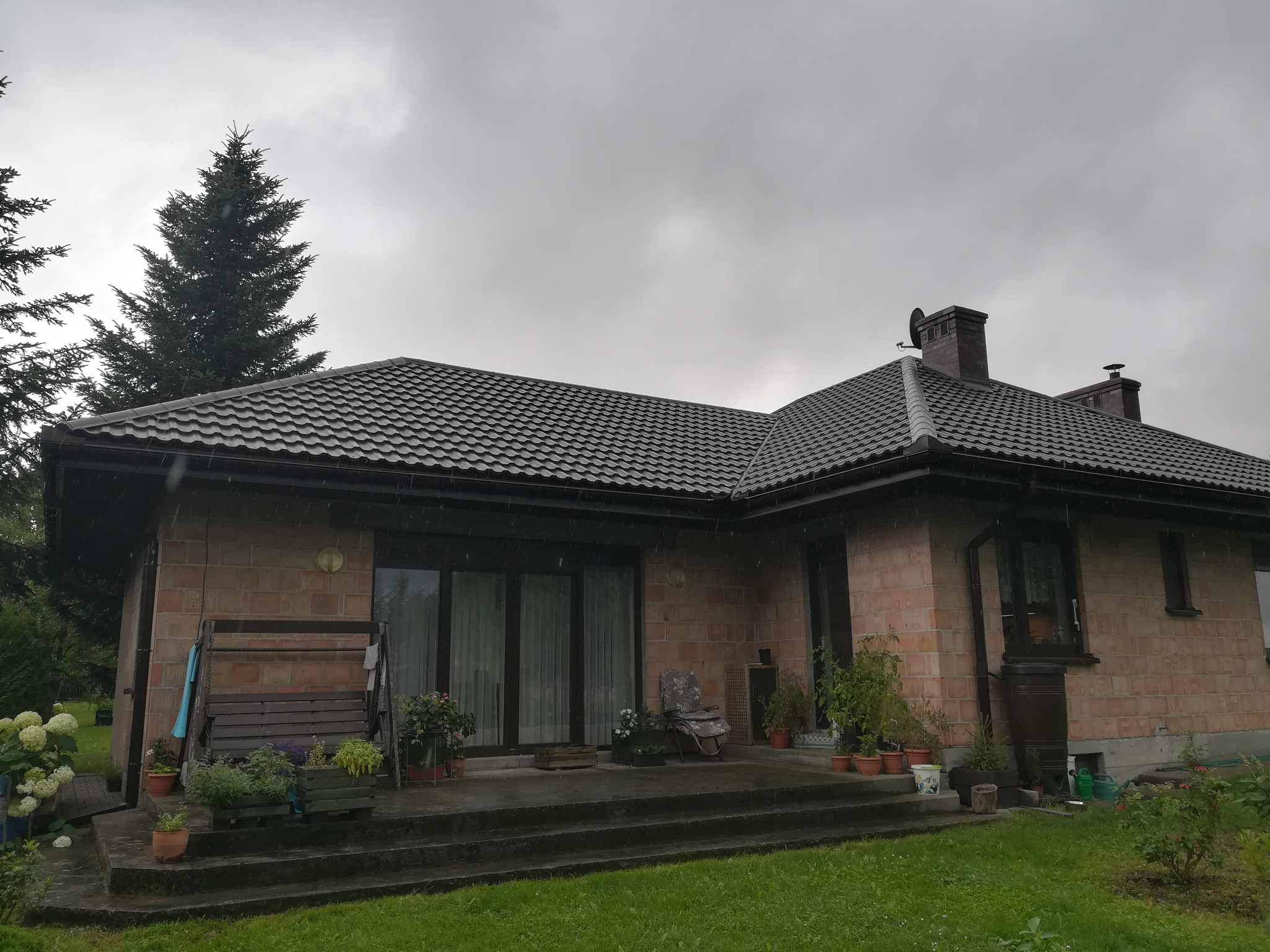 Dom jednorodzinny z brązową dachówką ceramiczną w pochmurny dzień, widoczna część elewacji z czerwonej cegły, roślinność w donicach na tarasie.
