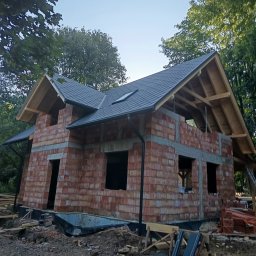 Usługi Budowlane - Surowy stan domu z czerwonej cegły z zamontowanym ciemnoszarym dachem i drewnianą więźbą dachową, otoczony zielenią drzew w słoneczny dzień.