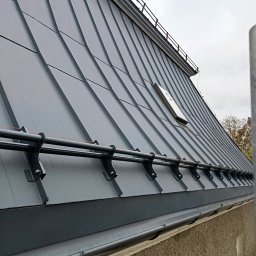 Usługi Budowlane - Szary dach z blachy trapezowej z oknem dachowym i systemem przeciwśniegowym, widok z dołu, na tle pochmurnego nieba.