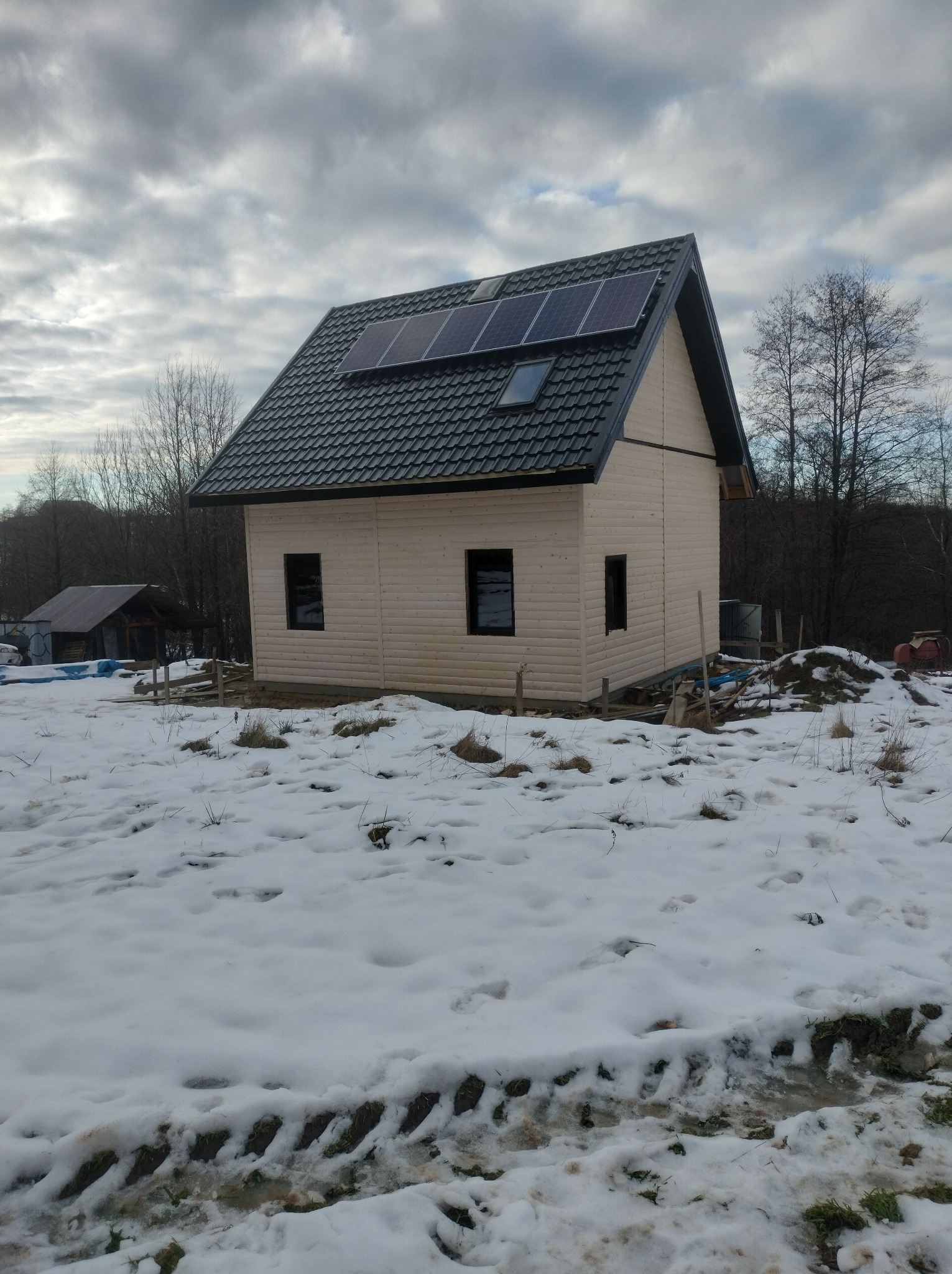 Niedokończony, drewniany dom z ciemnym dachem i panelami słonecznymi w zimowym krajobrazie. Widoczne czarne okna i częściowe ośnieżenie.