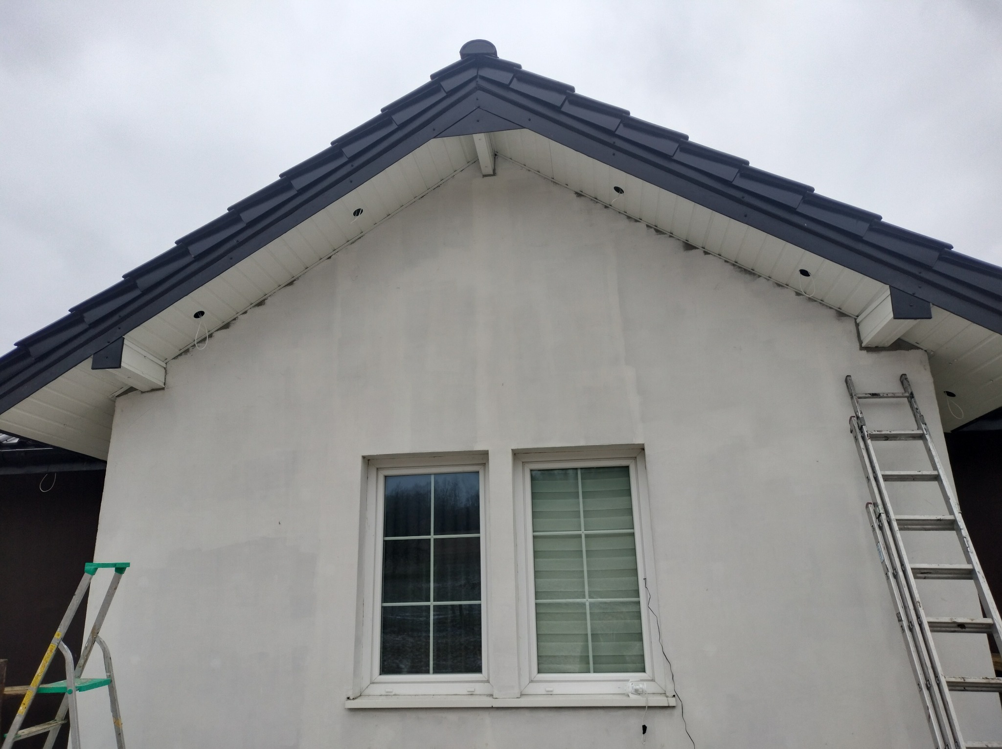Fragment domu z ciemną dachówką, białą elewacją i dwoma oknami. Przy ścianie oparta drabina. Widoczne otwory na oświetlenie pod dachem.