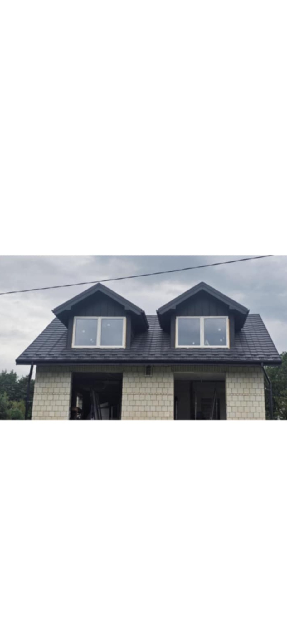 Dom w trakcie budowy z ciemnym dachem i dwoma oknami dachowymi. Ściany z jasnych bloczków, widoczne elementy konstrukcyjne w otworach okiennych.