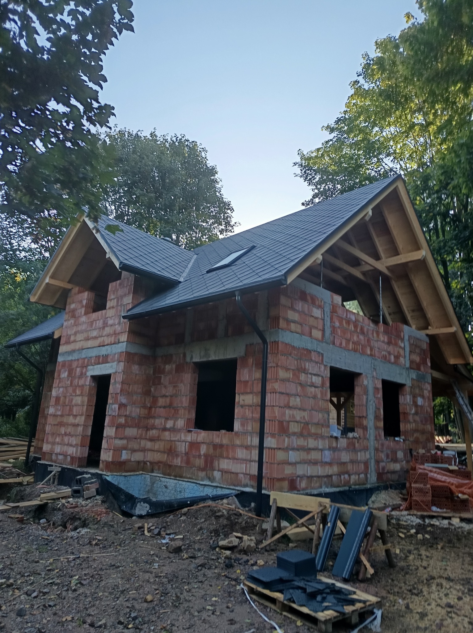 Surowy stan domu z czerwonej cegły z zamontowanym ciemnoszarym dachem i drewnianą więźbą dachową, otoczony zielenią drzew w słoneczny dzień.
