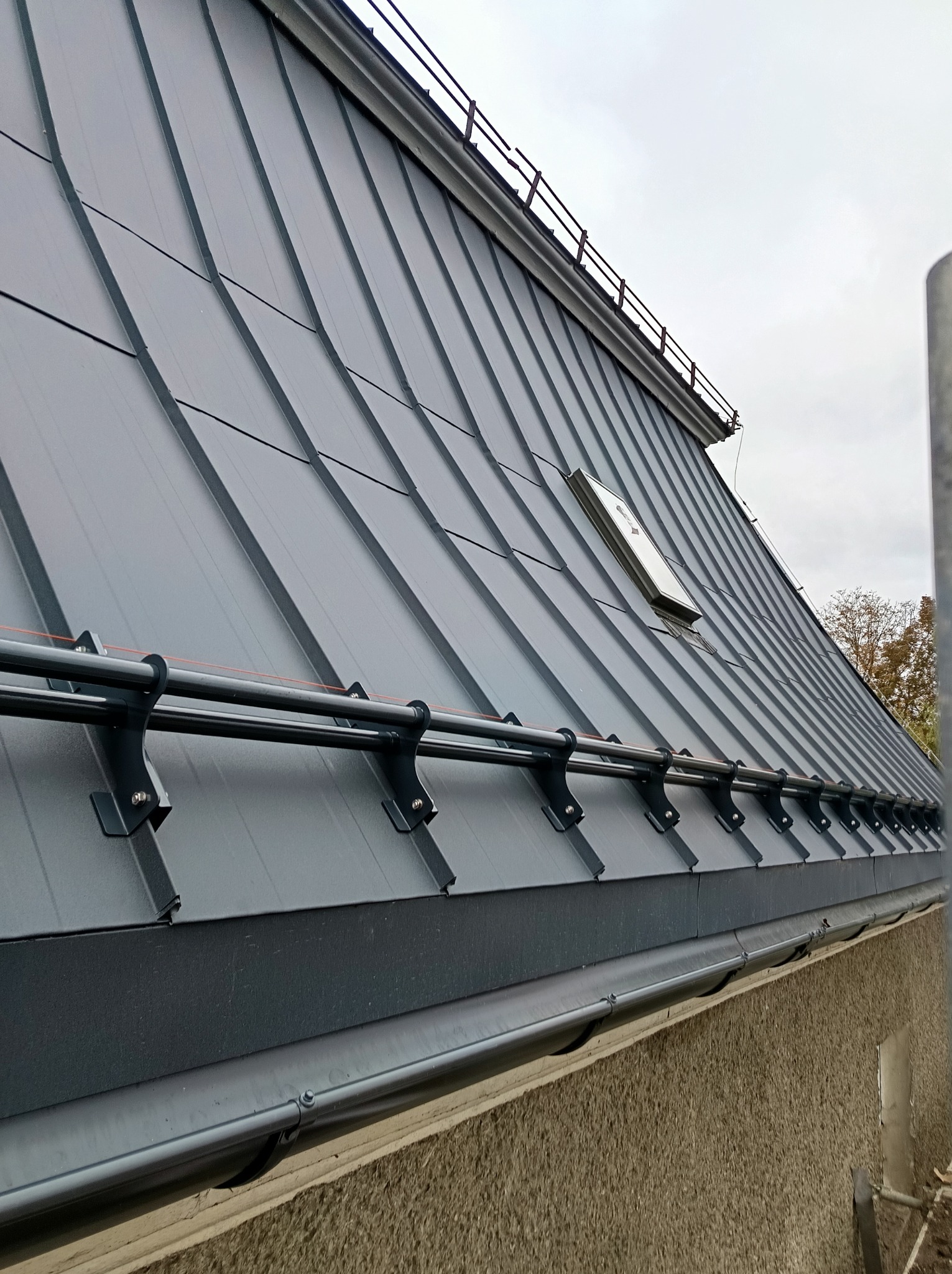 Szary dach z blachy trapezowej z oknem dachowym i systemem przeciwśniegowym, widok z dołu, na tle pochmurnego nieba.