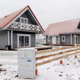 Dwa identyczne, szare domki z czerwonymi dachami i białymi balustradami, otoczone białym płotem na zaśnieżonym terenie, z widoczną skrzynką elektryczną na pierwszym planie.