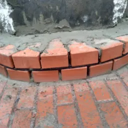 Obróbka podstawy komina z cegły klinkierowej, widoczne świeże spoiny i fragment muru kominowego w szarej zaprawie.