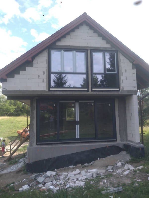 Surowy stan domu w trakcie budowy: ściany z białego bloczka, duże okna z czarnymi ramami, widoczne elementy konstrukcyjne, gruz budowlany na zewnątrz, teren wokół domu w trakcie prac ziemnych.