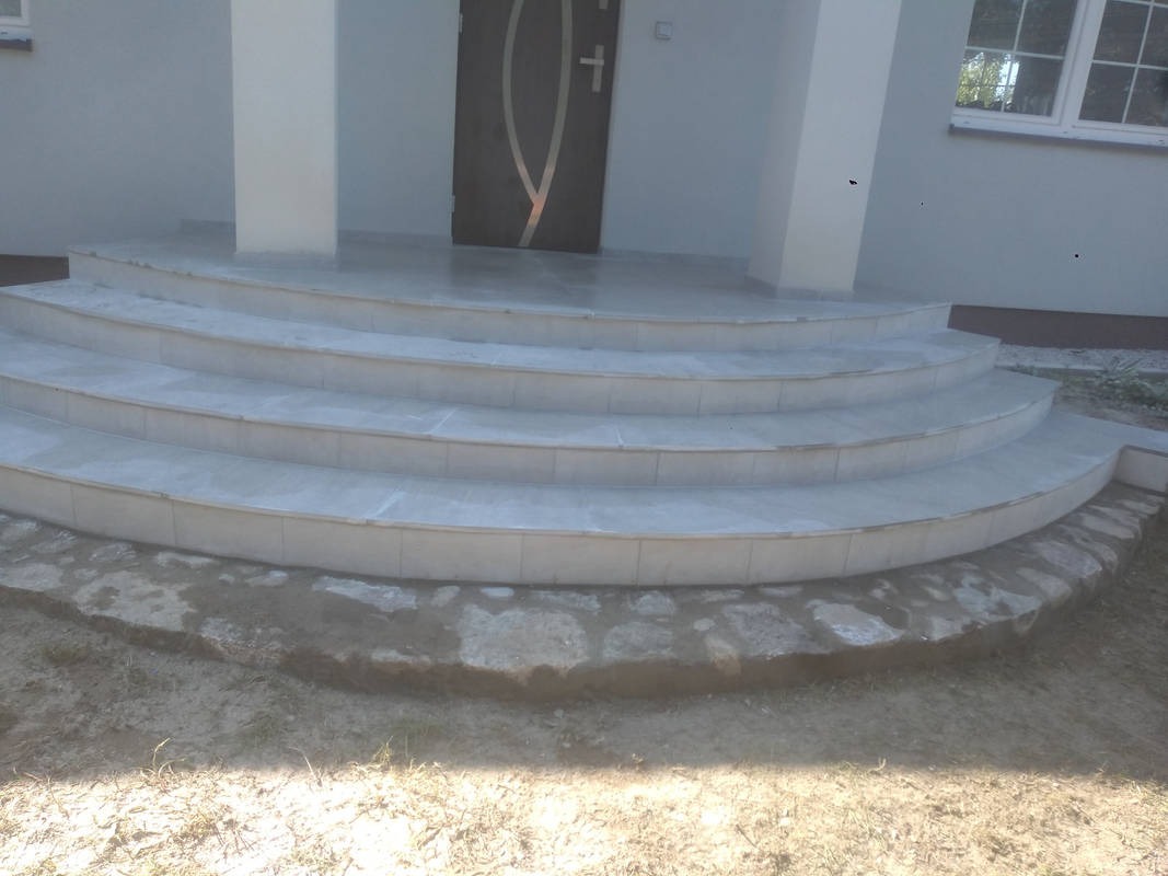 Wykończone, półokrągłe schody wejściowe do domu, wyłożone jasnymi płytkami ceramicznymi, z widocznym fragmentem elewacji i stolarki okiennej.