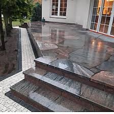 Wykończony taras z nieregularnych płyt kamiennych w odcieniach szarości i brązu, prowadzący do białego budynku z dużymi oknami, widok z góry na schody i ścieżkę obok tarasu.