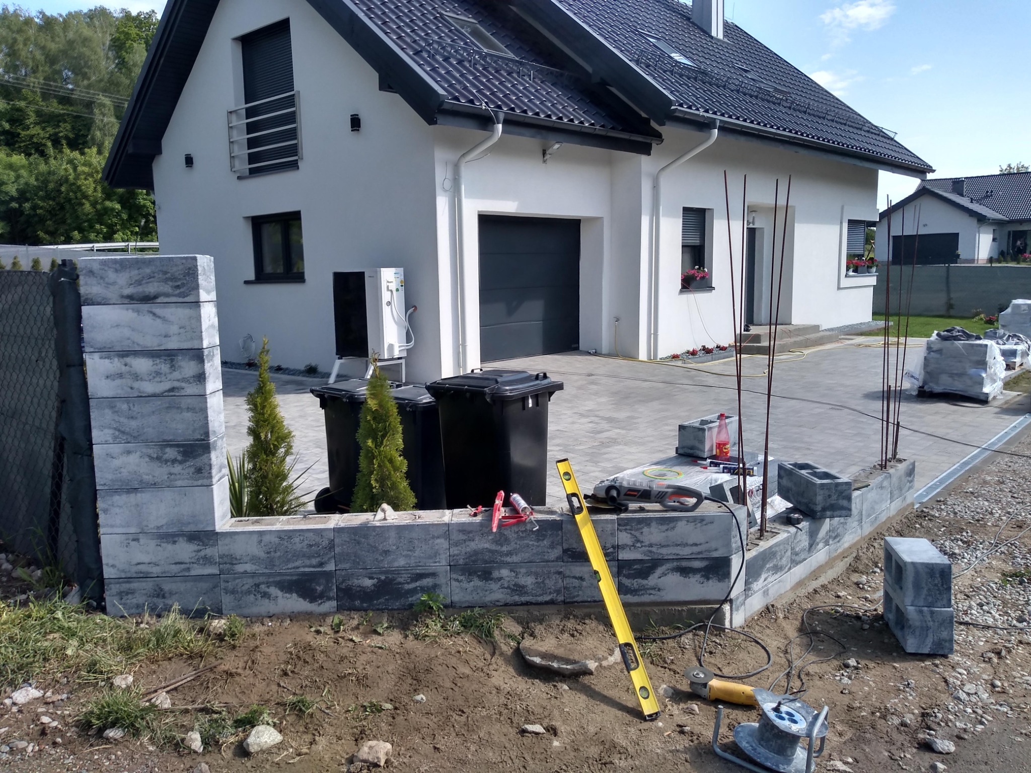 Budowa ogrodzenia z szarych bloczków betonowych na tle nowoczesnego domu z białą elewacją i ciemną dachówką; widoczne narzędzia budowlane, poziomica, oraz elementy przygotowane do dalszej pracy.